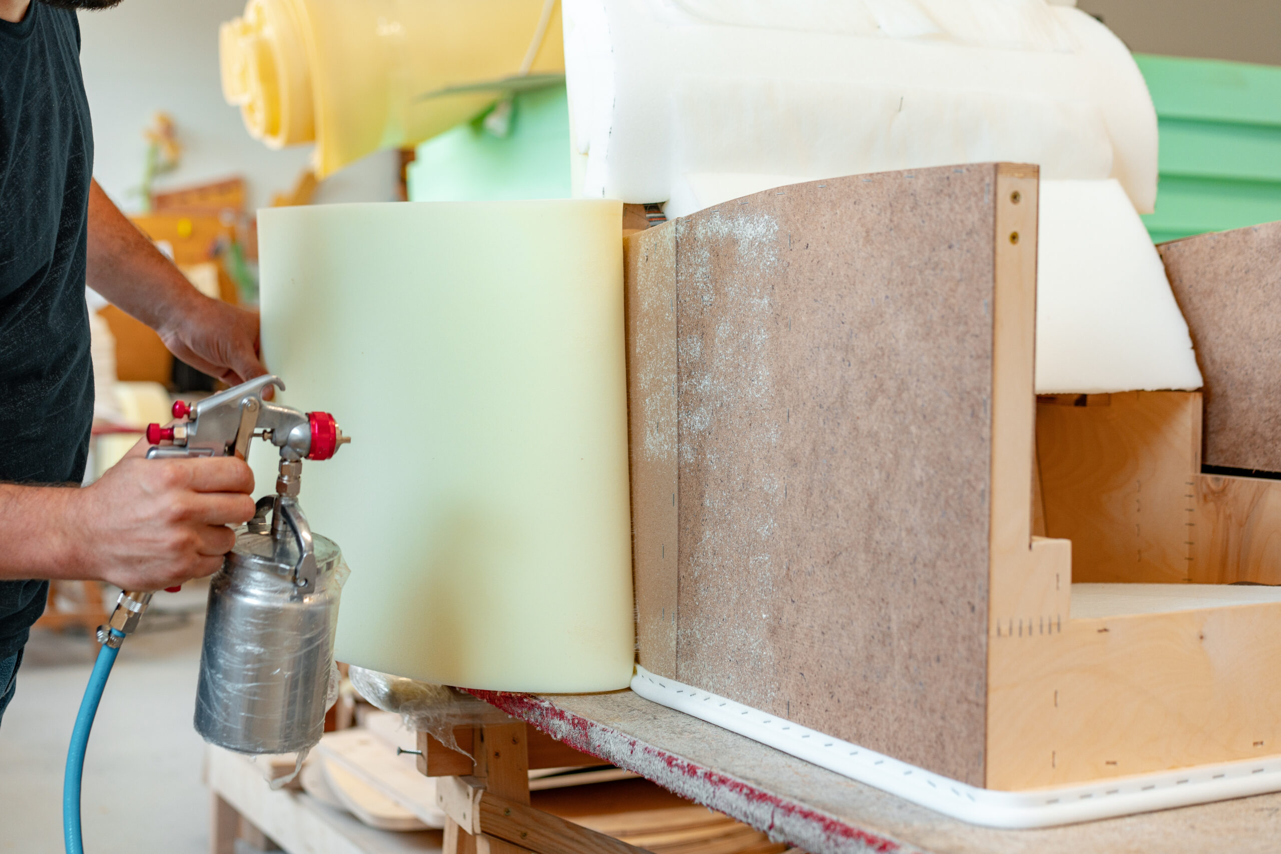Person applying adhesive spray to foam and wood for upholstery work.
