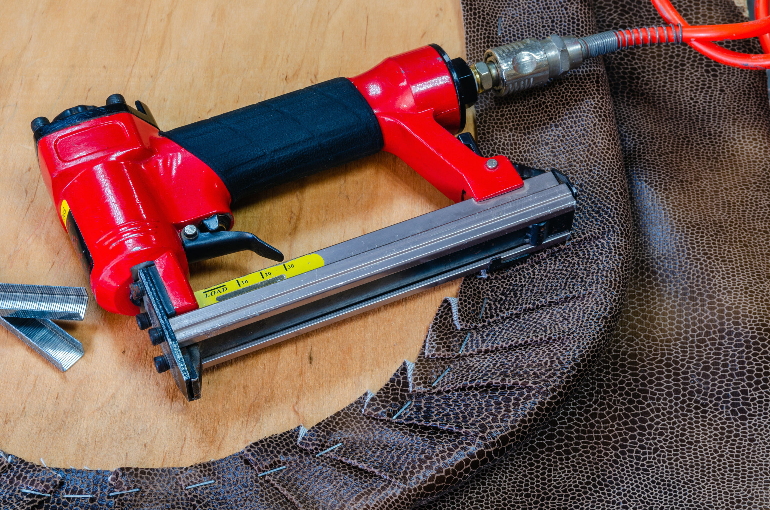 Red pneumatic stapler with staples and upholstery fabric on wooden surface.