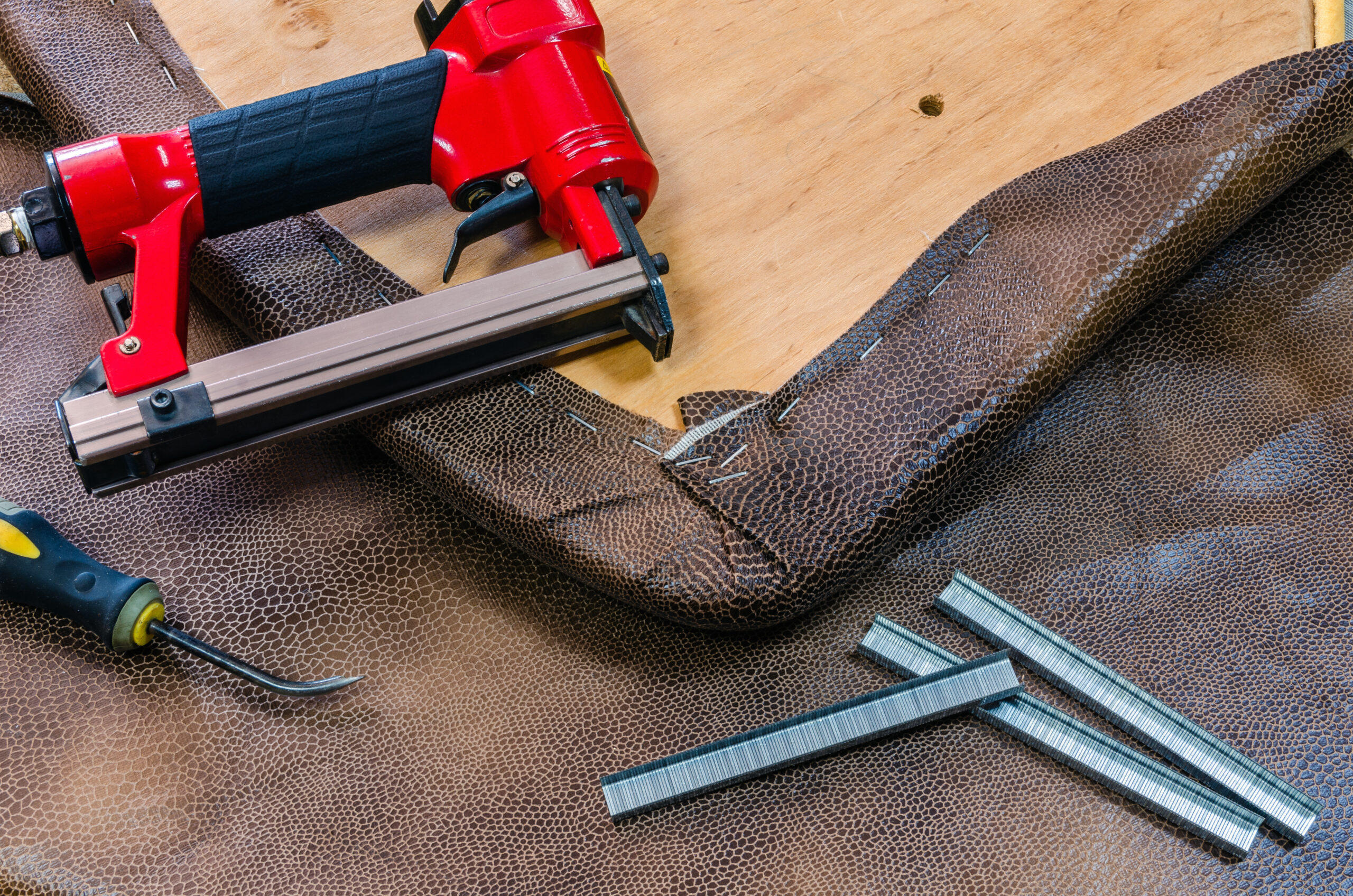 Upholstery stapler and tools on brown fabric with staples nearby, for DIY furniture repair.
